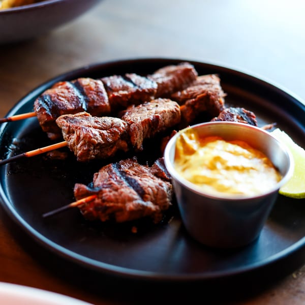 Grilled beef skewers with a side of mustard sauce and a lime wedge on a black plate.