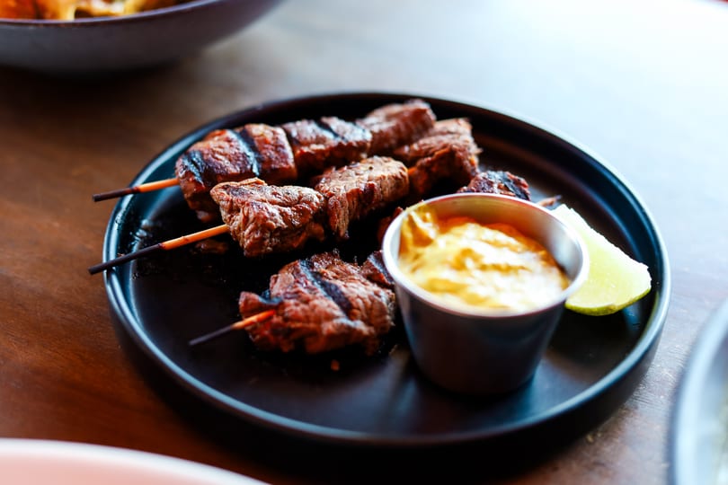 Grilled beef skewers with a side of mustard sauce and a lime wedge on a black plate.