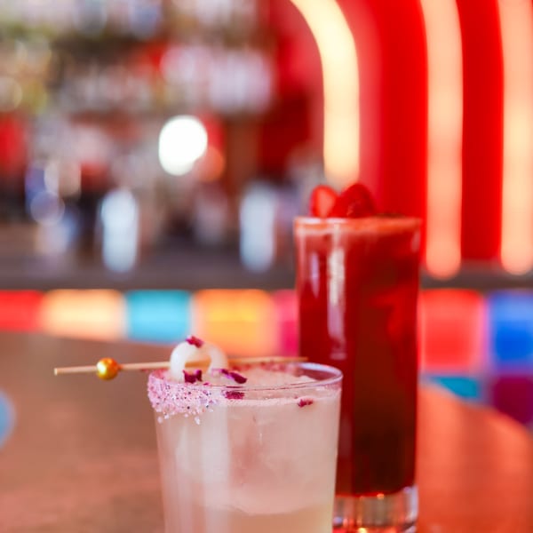 Two vibrant cocktails on a bar, one light with a lychee garnish, the other dark red.