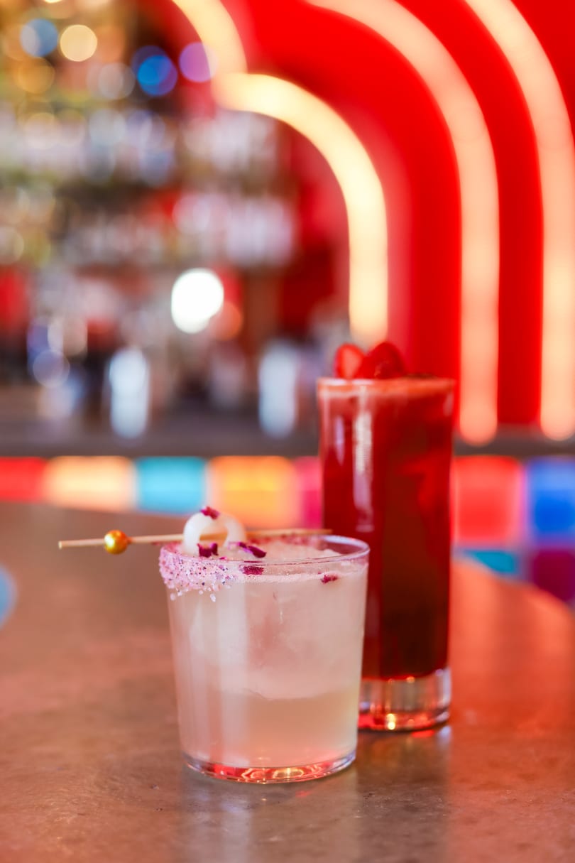 Two vibrant cocktails on a bar, one light with a lychee garnish, the other dark red.