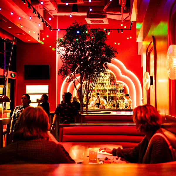 A vibrant, red-lit bar interior with patrons, a tree, and a well-stocked bar with bartenders.
