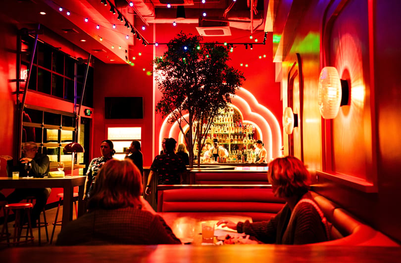 A vibrant, red-lit bar interior with patrons, a tree, and a well-stocked bar with bartenders.
