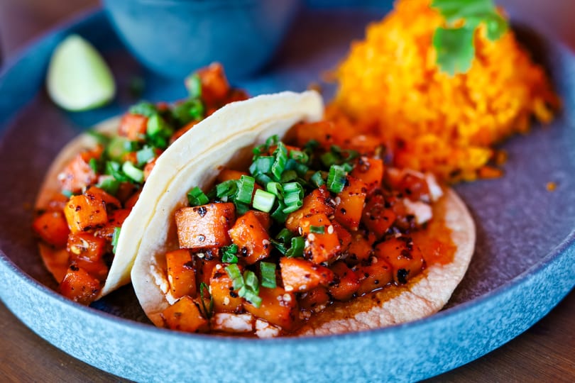 Two tacos filled with diced sweet potatoes and green onions, served with orange rice and lime.