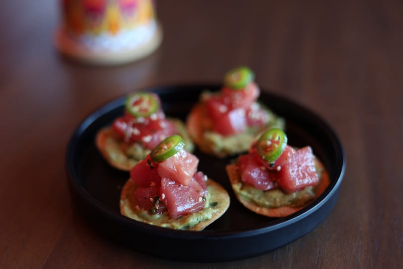 Four tuna poke appetizers with avocado and jalapeño slices on a black plate.