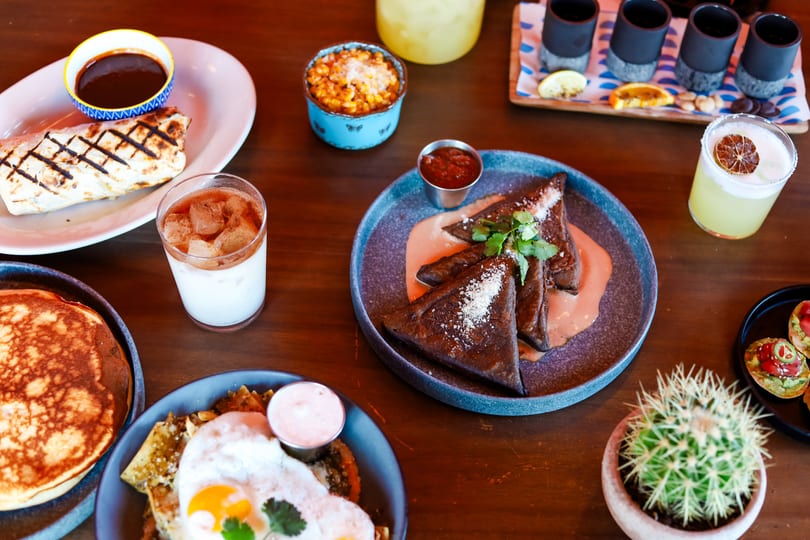A table laden with various Mexican dishes, drinks, and a small potted cactus.