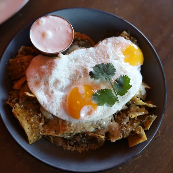 Chilaquiles verdes with two fried eggs, cilantro, and a side of crema in a dark bowl.