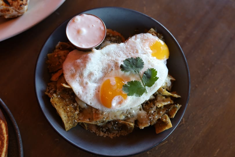 Chilaquiles verdes with two fried eggs, cilantro, and a side of crema in a dark bowl.