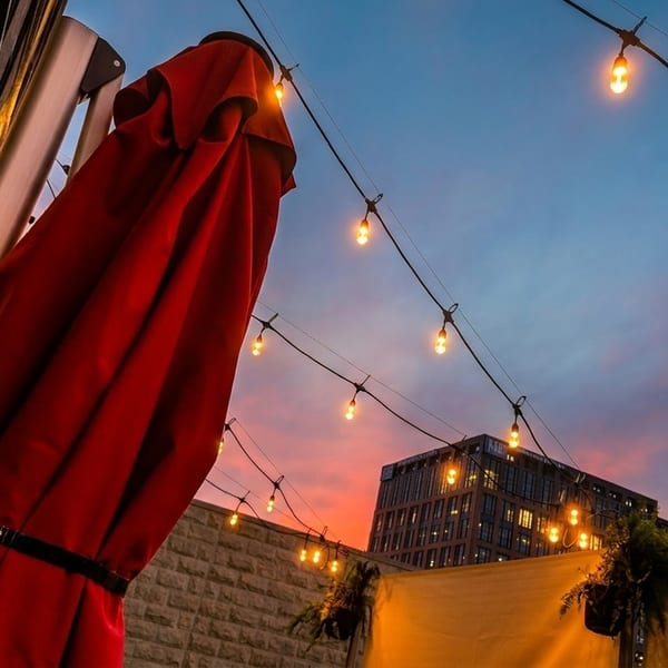 Red patio umbrella and string lights against a vibrant sunset sky with a city building.