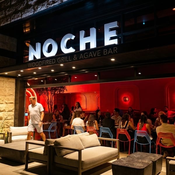 Noche Woodfired Grill & Agave Bar exterior at night, with patrons dining inside and lounge seating outside.