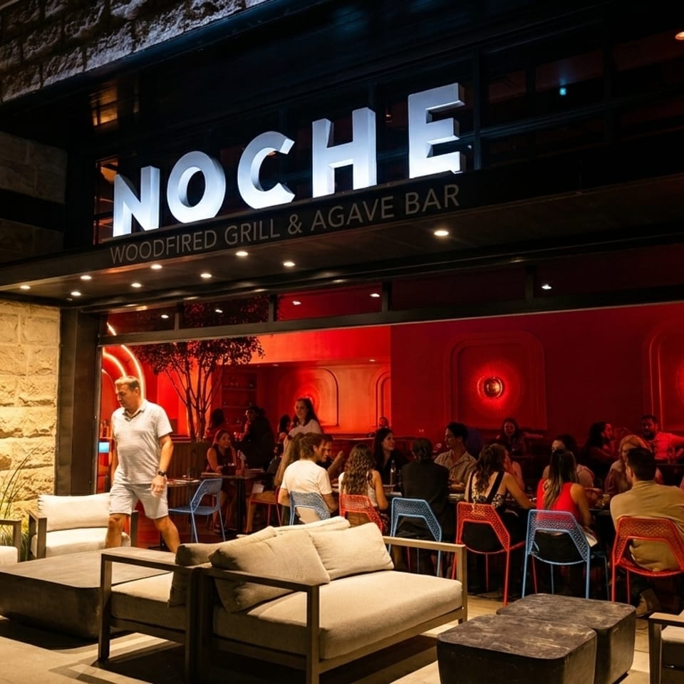 Noche Woodfired Grill & Agave Bar exterior at night, with patrons dining inside and lounge seating outside.
