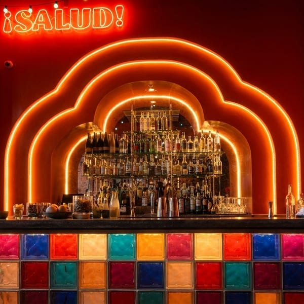 Vibrant bar with a colorful glass block counter, neon "¡SALUD!" sign, and arched lighting.