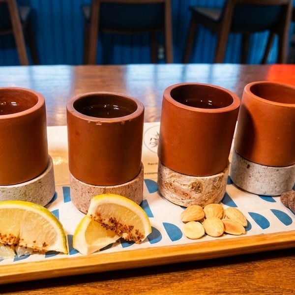 Four clay cups with drinks, lemon slices, almonds, and a chocolate on a wooden tray.