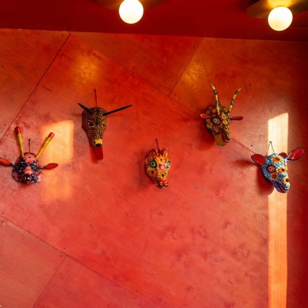 Colorful animal masks adorn a vibrant red wall, illuminated by warm sunlight and ceiling lights.